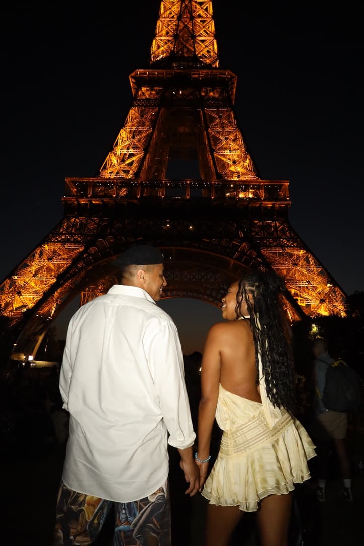 Cayla and Shaddy hold hands and look at each other beneath the illuminated Eiffel Tower.