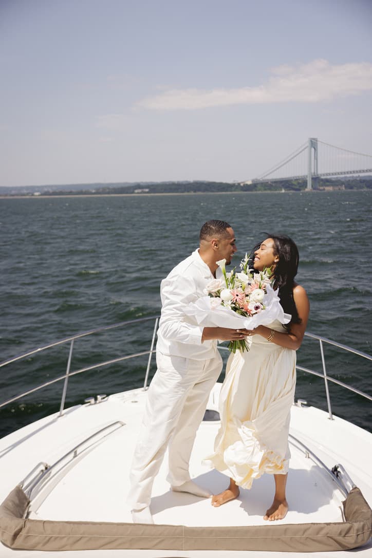 Cayla and Shaddy laughing together with a bouquet on the bow of a boat.